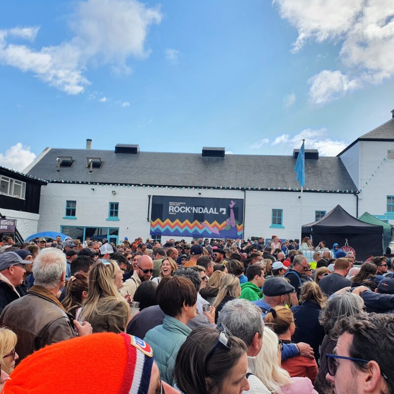 A crowd of people laughing and smiling and drinking with a large Distillery building in the background