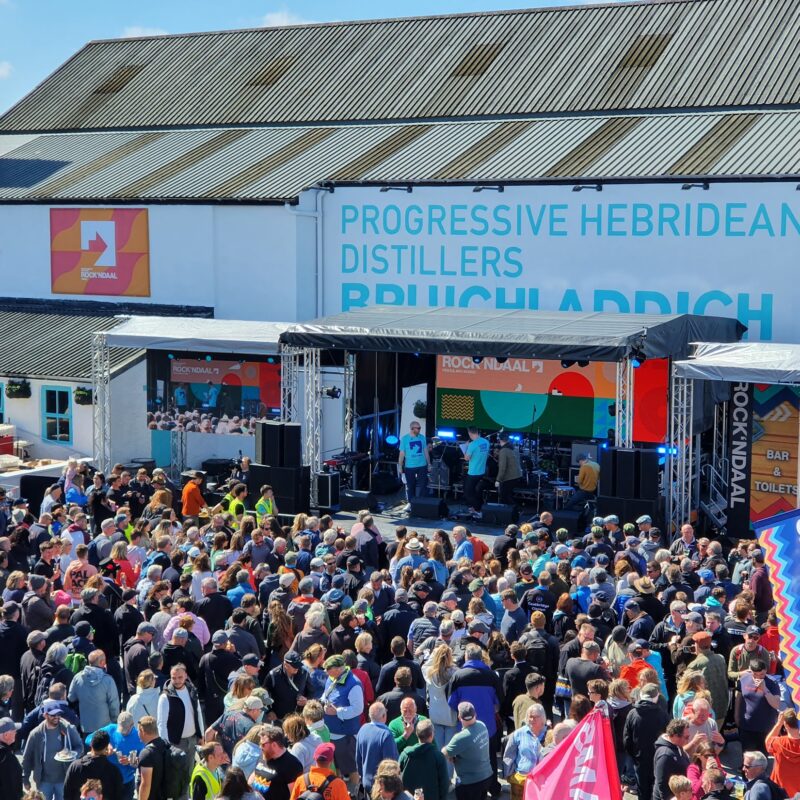 A large crowd of people in front of a stage with a band performing on stage. A large distillery building behind the stage.