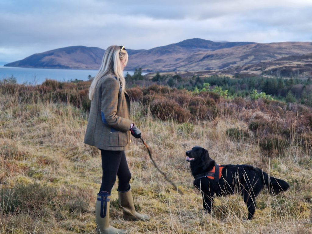 Girl throwing stick on hillside, with dog beside her. Sea in background.