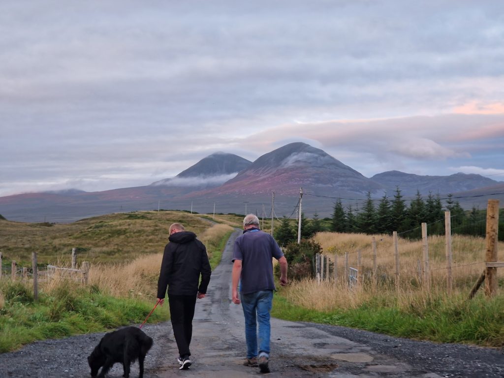 Two men walking along a road with a dog. Hills in background