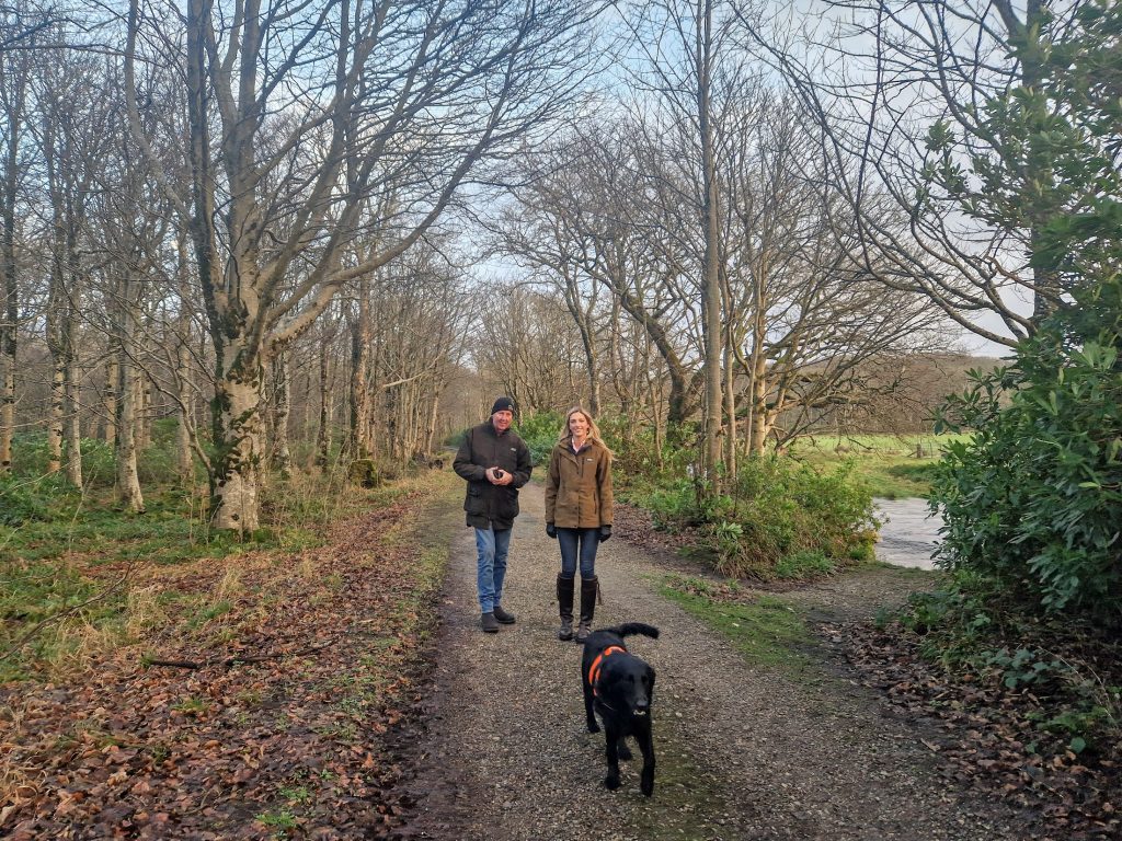 Two people stood on track in woods beside a river. Dog walking towards them