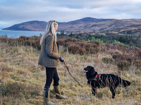 A girl holding a stick and a dog watching the stick. Hills and sea in the background