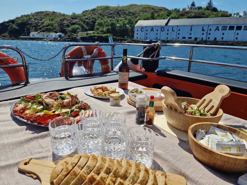 A table laden with seafood on a boat, with the sea and Caol ila Distillery in the background