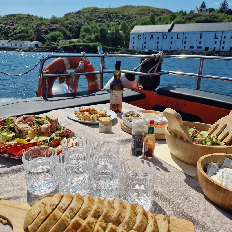 A table laden with seafood on a boat, with the sea and Caol ila Distillery in the background