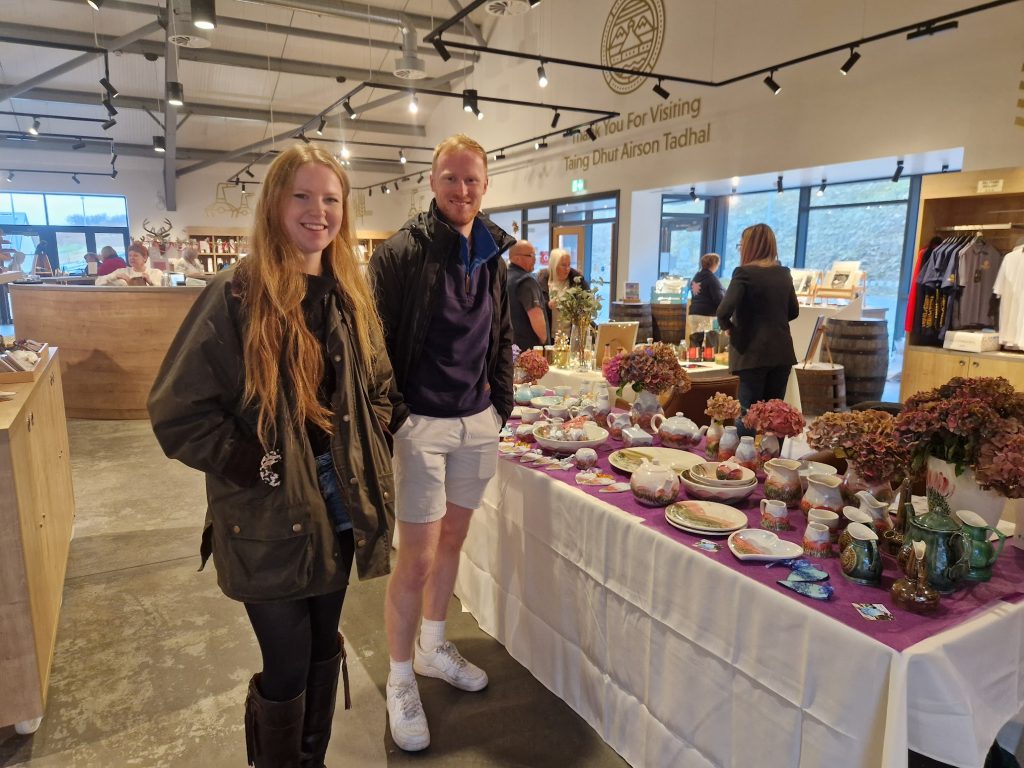 Two people stood smiling beside a table filled with ceramics.