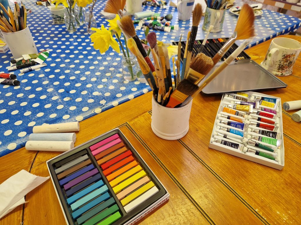 Pastel crayons and paints on a table with a mug of brushes