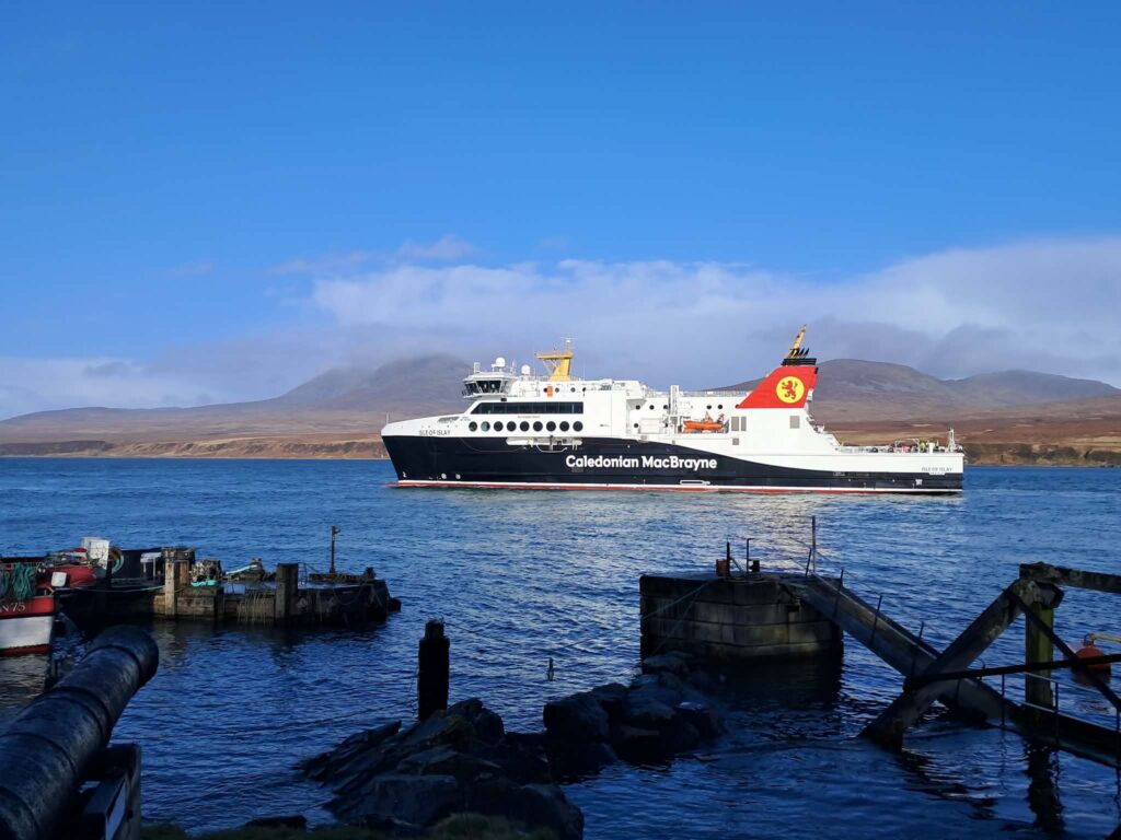 A large ferry boat on the sea with a jetty in the foreground. Hills in the background